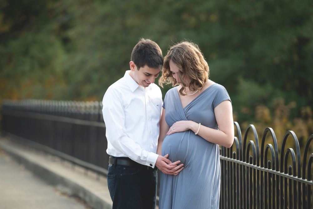 A couple affectionately looking at the woman's pregnant belly while standing on a bridge.