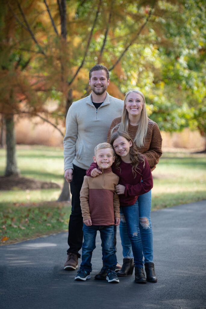 A smiling family of four poses for a photo on a tree-lined path.