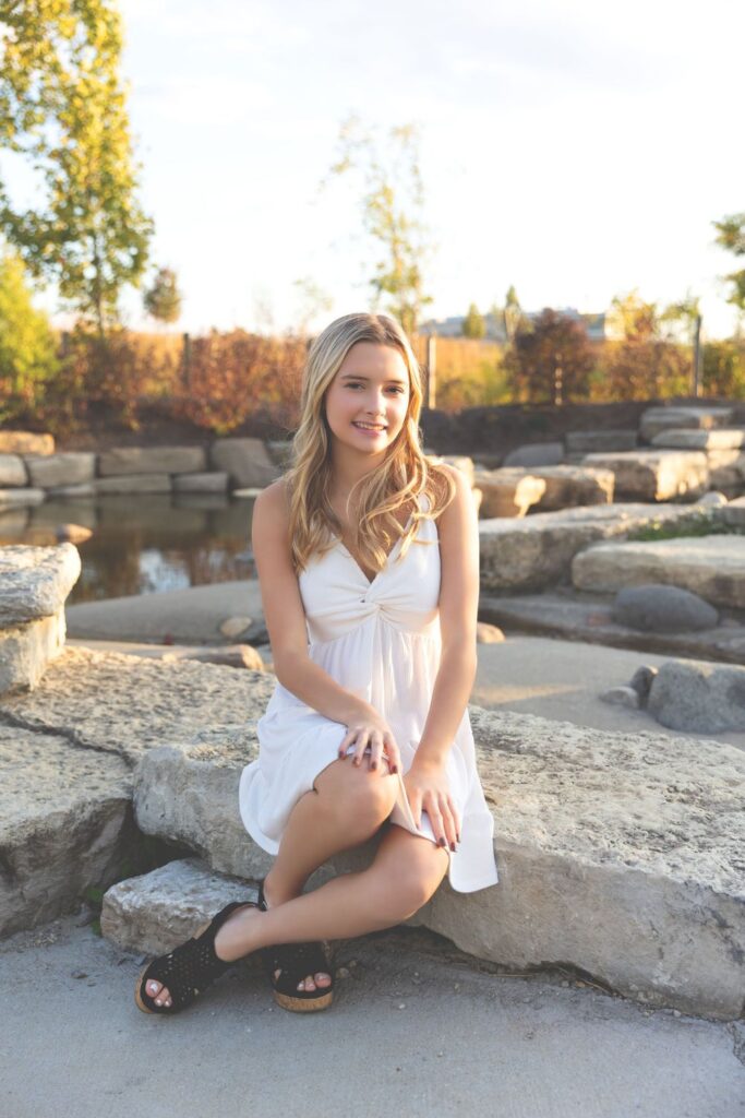 A woman sitting on a rock by a pond in a white dress at sunset.