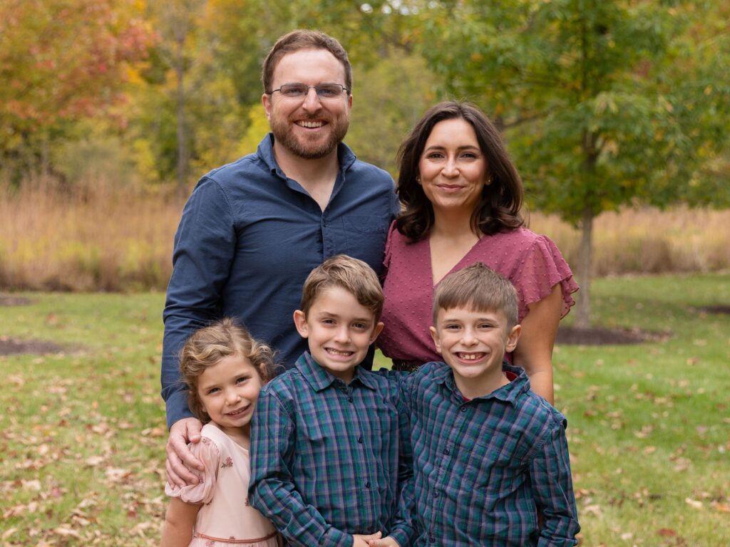 A family portrait of two adults and three children smiling outdoors with autumn foliage in the background.