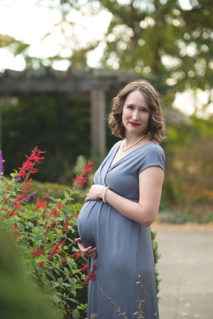 A pregnant woman smiling gently while standing beside flowering plants in a garden.