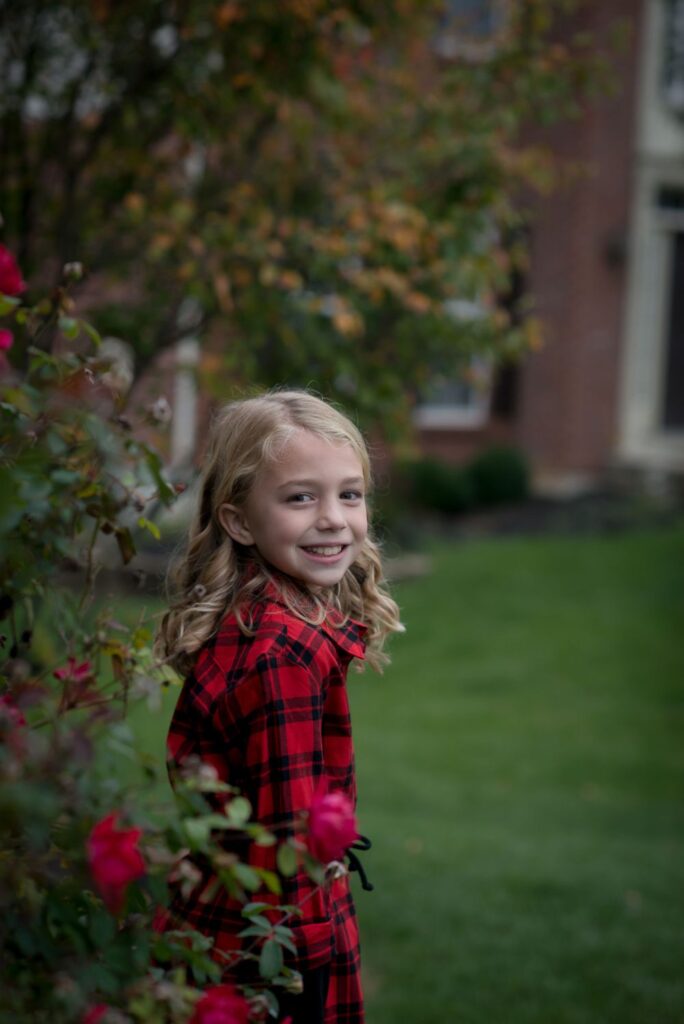 A smiling young girl in a red plaid dress standing beside rose bushes.