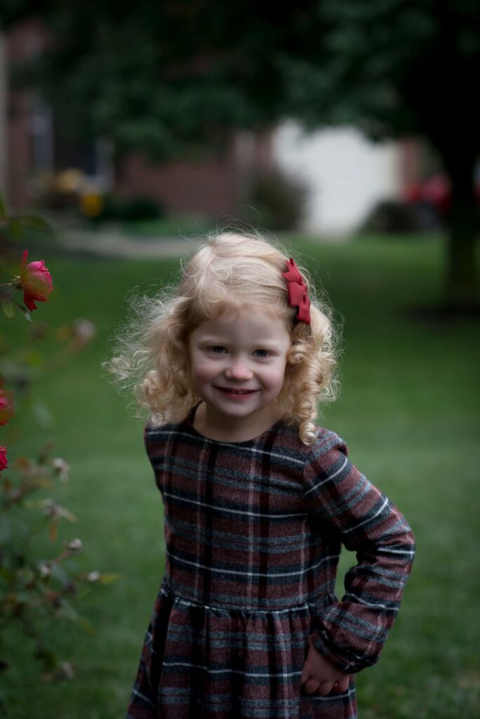 A young girl with curly hair, wearing a plaid dress and a red hair clip, smiling in a garden setting.