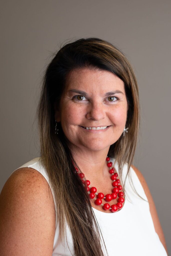 Professional headshot of a smiling woman with long hair, wearing a white sleeveless top and a red necklace.