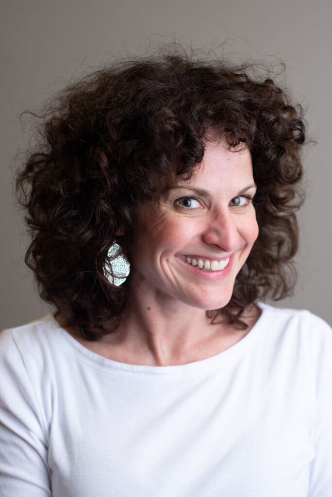 A woman with curly hair smiling at the camera, wearing a white top and silver earrings.