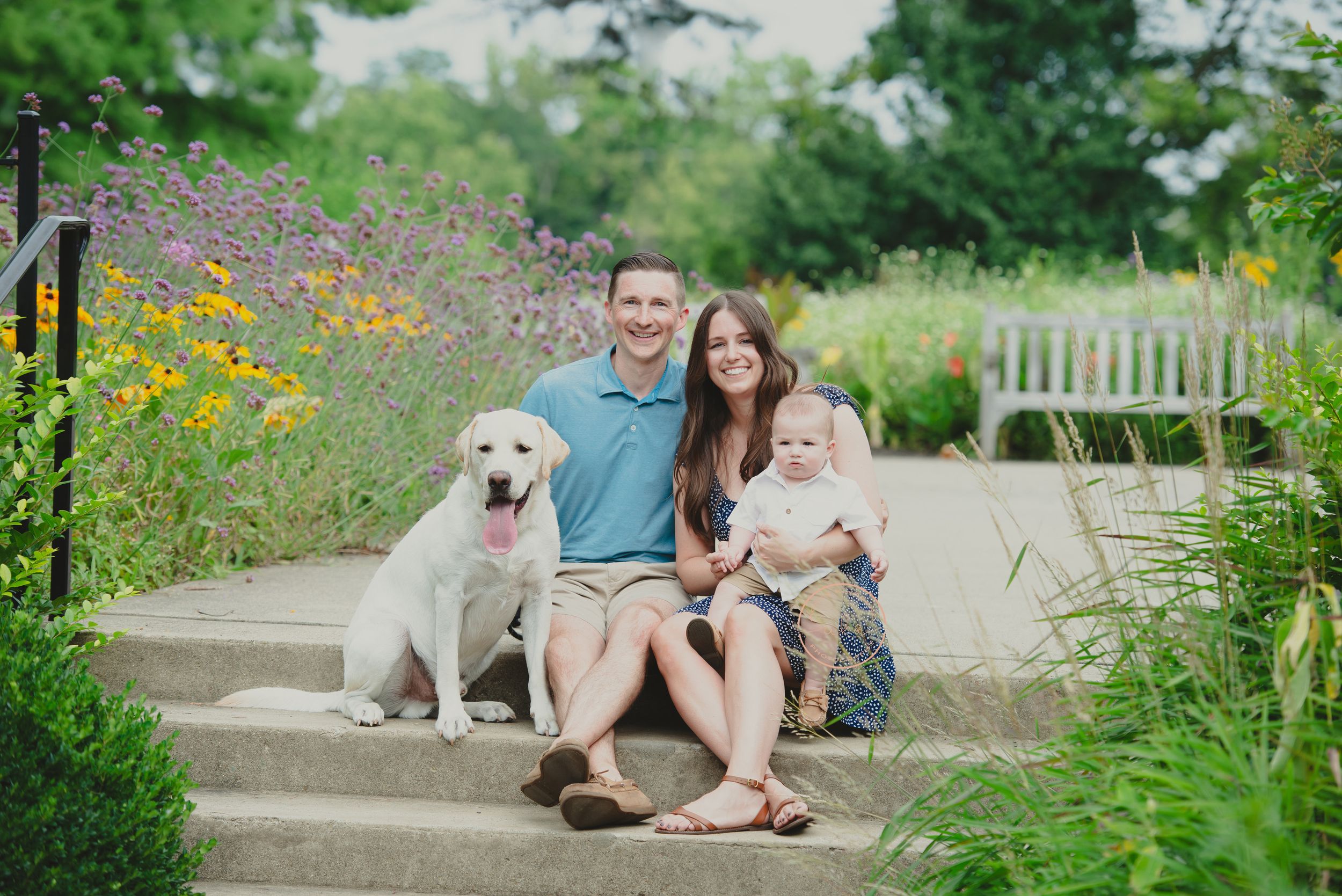 A family portrait on outdoor steps featuring a man, a woman holding a baby, and a dog surrounded by greenery and flowers.