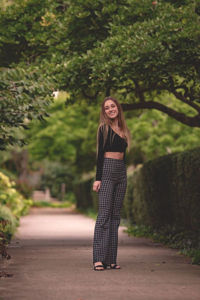 A woman smiling as she stands on a garden path lined with lush green hedges.