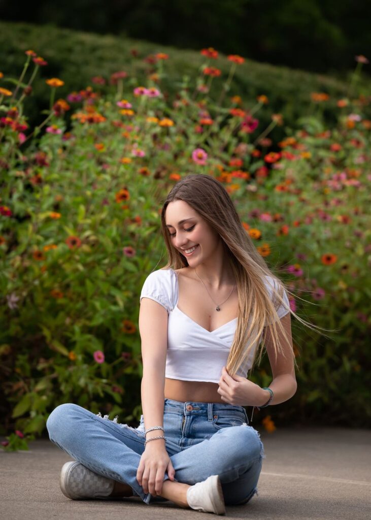 A young woman sitting cross-legged in front of a flowerbed, smiling.