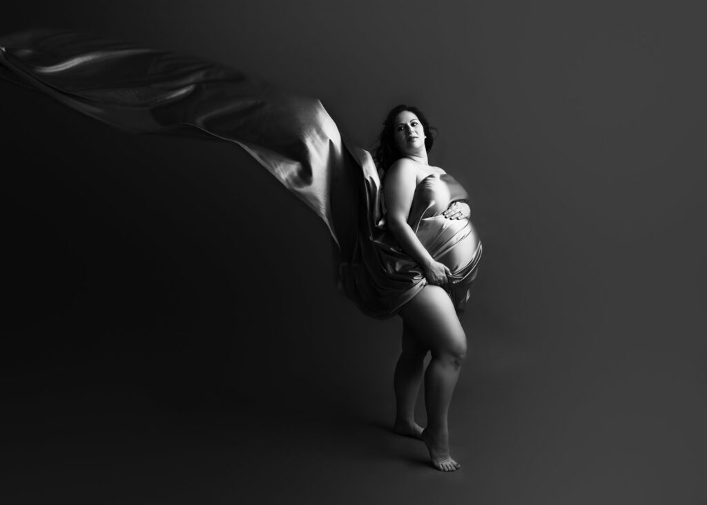 A monochromatic image capturing a pregnant woman draped in flowing fabric, posing with a sense of serenity.