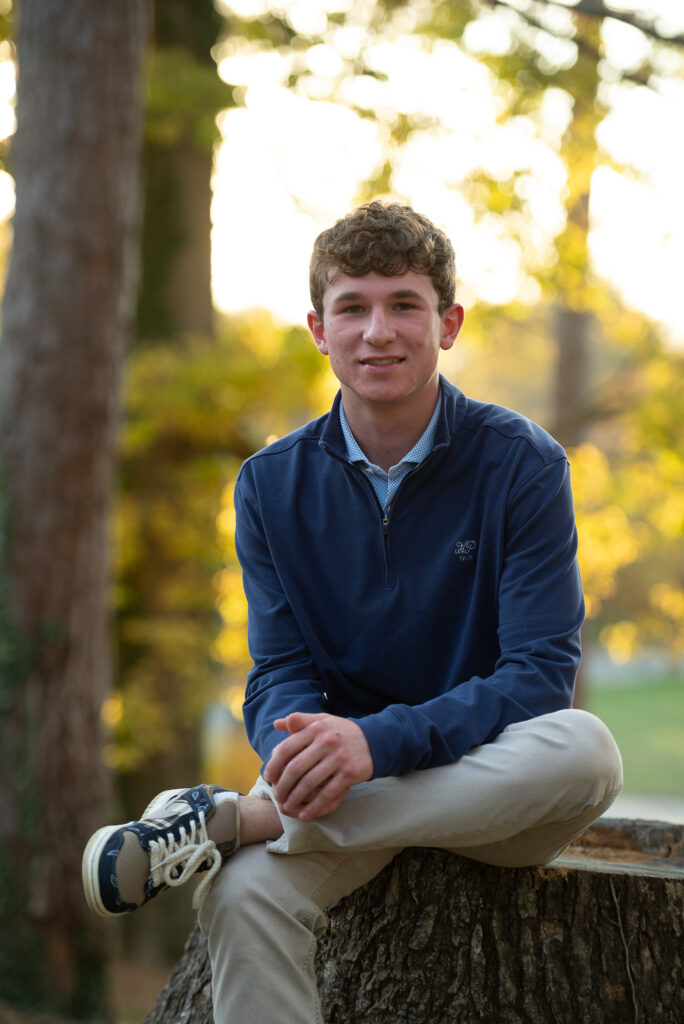 A young man sitting on a tree stump with autumn trees in the background.