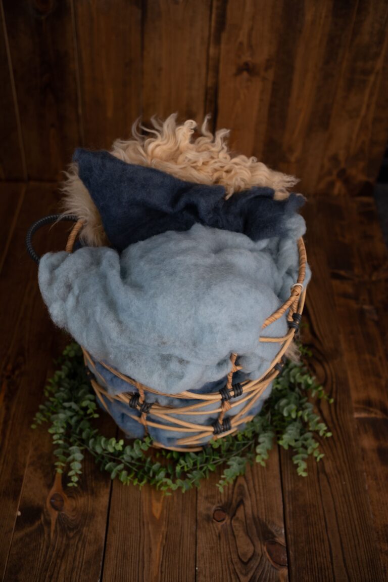 Wicker basket with blue and grey blankets, an infertility awareness sheepskin rug, placed on a wooden background and surrounded by greenery.