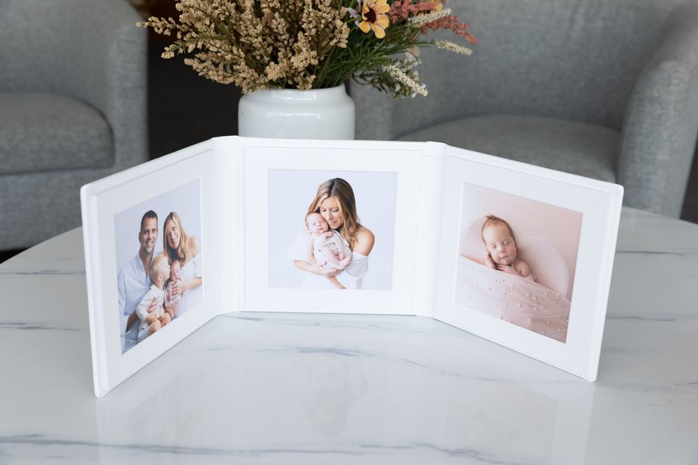 A white tri-fold picture frame displaying family photographs on a marble table, with a vase of flowers in the background.