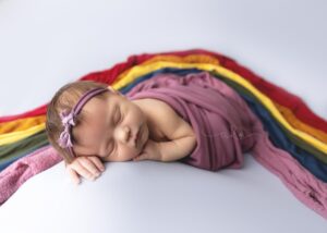 Newborn baby sleeping peacefully on colorful fabrics, swaddled in a purple cloth with a matching headband.