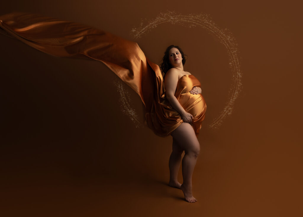 Pregnant woman with flowing silk fabric on brown backdrop.