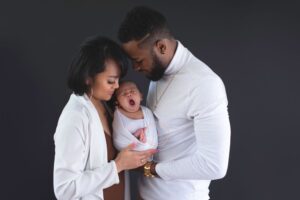 A couple tenderly holds their yawning baby against a plain background.