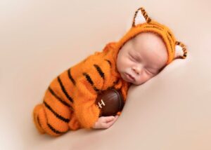 Newborn dressed in a tiger-striped outfit holding a small football, sleeping peacefully.