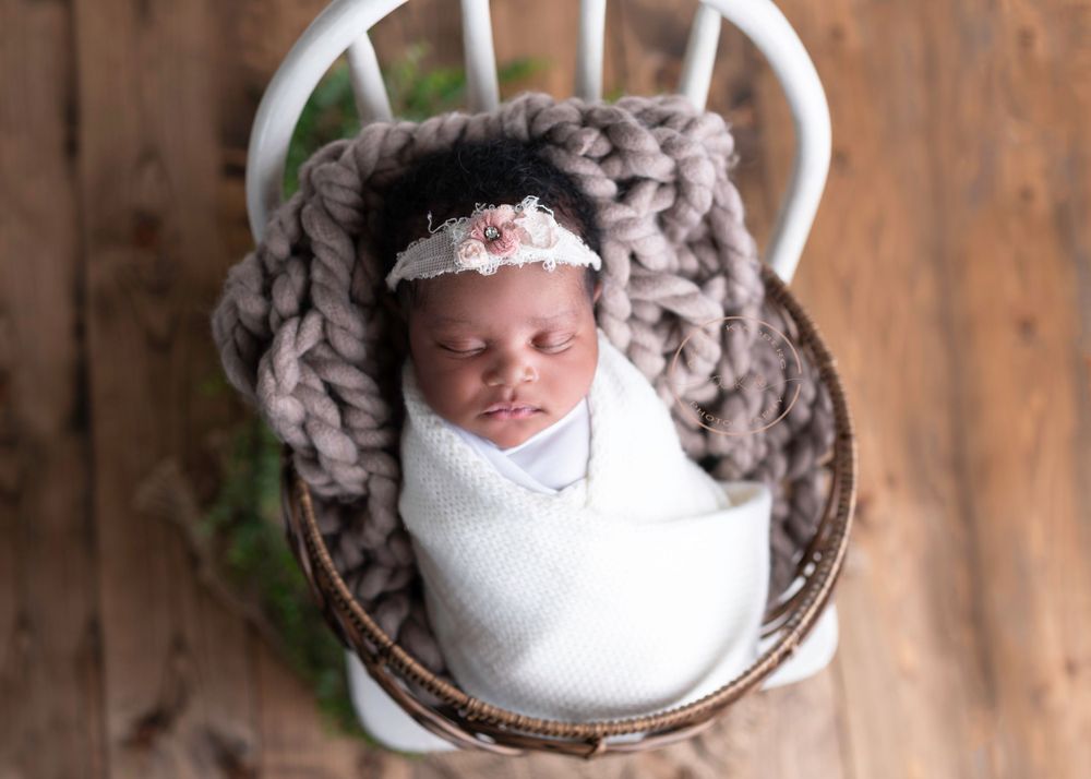 A newborn baby wrapped in a white blanket, resting peacefully in a basket with a chunky knit layer and adorned with a lace headband.