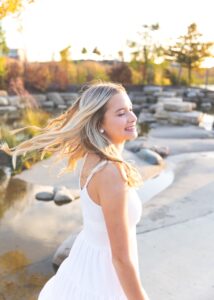 A high school senior girl in a white dress smiles joyfully, her blonde hair flowing, as she strolls by a serene lakeside park at sunset.