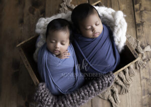 Two infants sleep peacefully, swaddled in blue blankets, and nestled side by side in a wooden crate. Positioned on a cozy knitted blanket on a wooden floor, they are surrounded by soft, chunky-knit fabrics in neutral tones—a perfect setting for newborn photography that captures warmth and serenity.