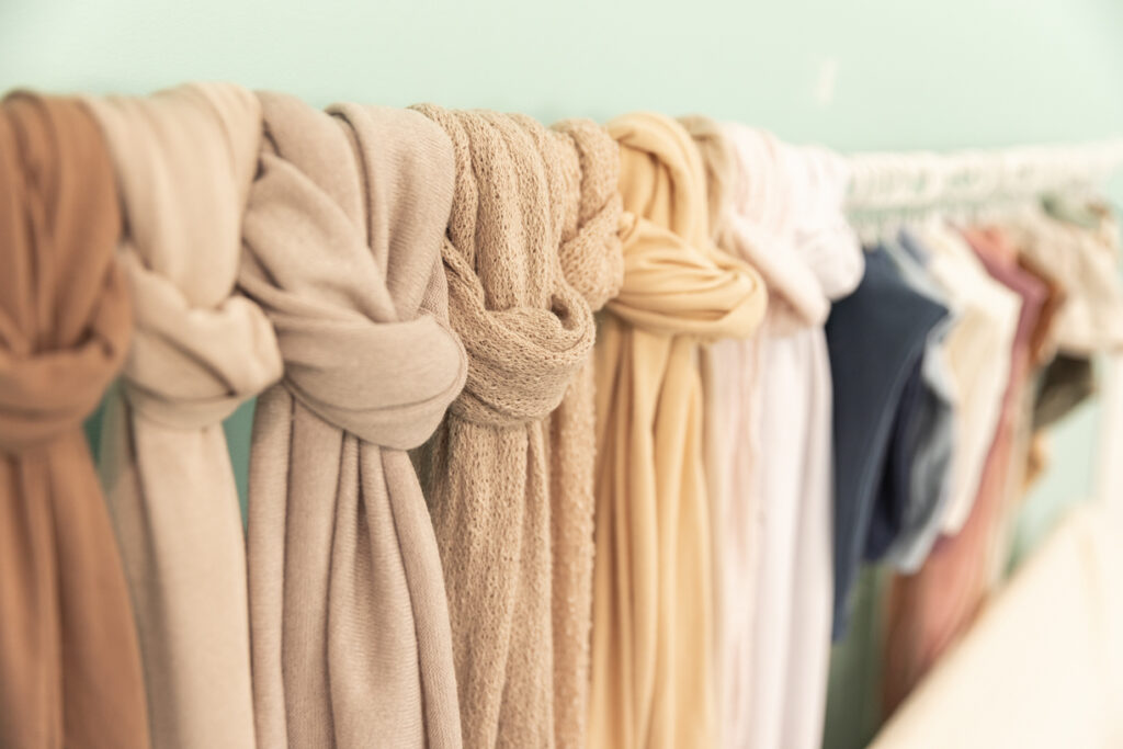 Close-up of a row of soft, neutral-toned scarves hanging on a rack against a light turquoise wall. The scarves are knotted at the top, creating a casual yet organized display—perfect for a fashion blog or an SEO feature. The colors range from beige to light brown to pastel hues.