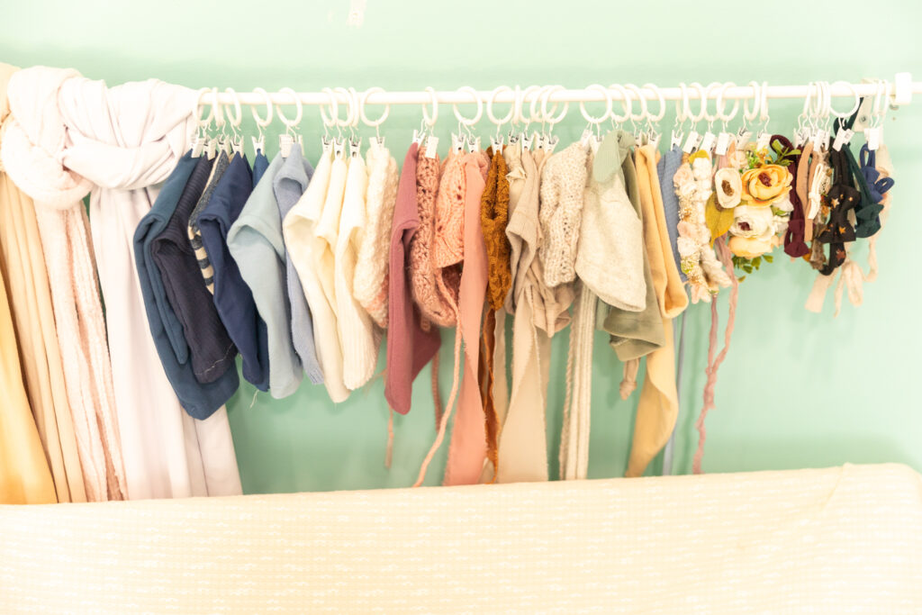 Various pastel-colored baby bonnets and hats are hanging on a white rack against a light green wall in the photo studio. A white blanket partially drapes over a hanging rod in the foreground, adding to the serene setup.