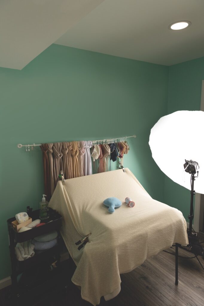 A photography studio setup with a teal green wall. A tabletop covered with a beige cloth is lit by a large light with a circular diffuser on the right. Various photography equipment, towels, and baby props are spread on and around the table, ready for SEO-friendly blog content creation.