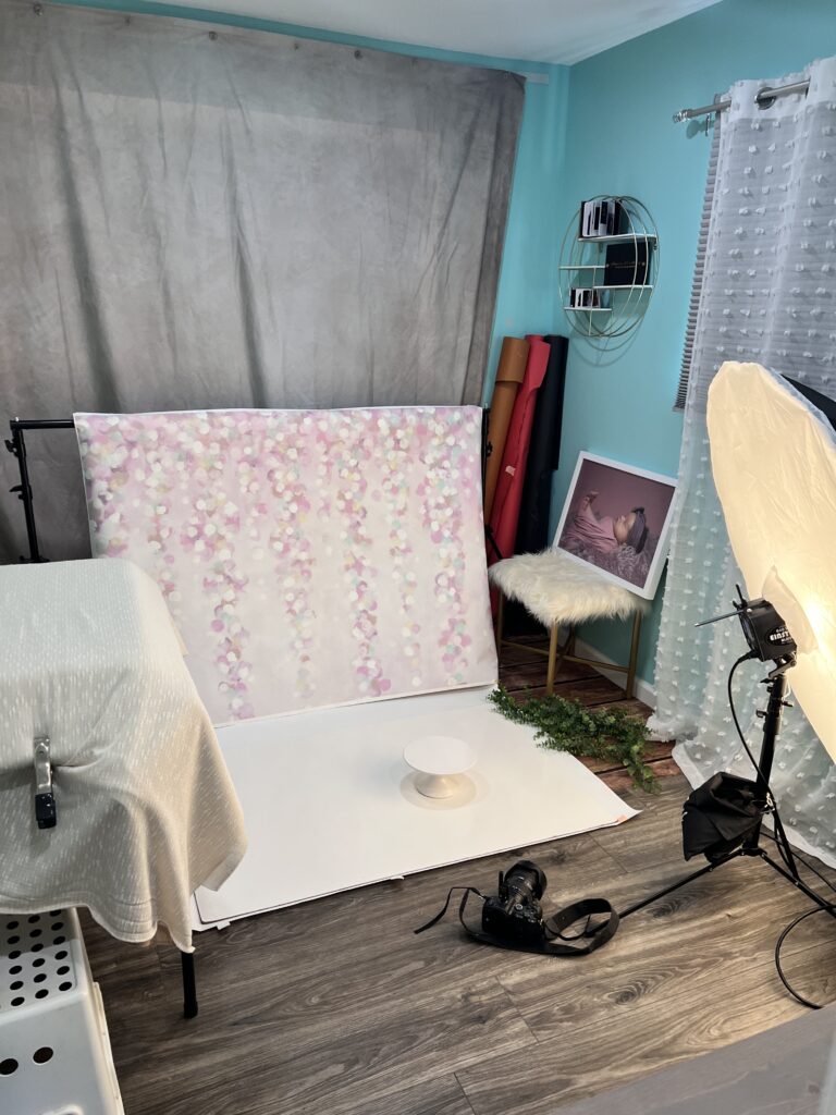 A photo studio setup for Cincinnati newborn photography features a backdrop of hanging pink and white flowers, arranged with softboxes, a camera on the floor, and a small white pedestal in the center. A chair with a blanket and a portrait on the wall are also in view.