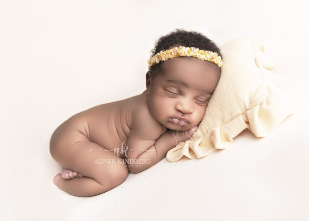 A newborn baby with closed eyes sleeps on a soft, cream-colored blanket, resting their head on a matching pillow. The baby wears a delicate beaded headband and lies on their side with legs slightly tucked in, capturing one of the sweetest posing ideas for newborn photography in a softly lit and minimalist background.