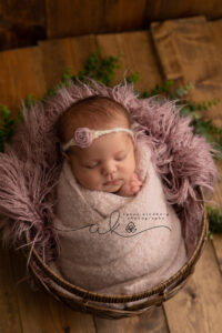 A newborn baby, swaddled in a soft pink blanket, sleeps peacefully in a round wicker basket. Ideal for newborn photography, the baby wears a headband adorned with a pink flower. The basket is cushioned with a fluffy mauve blanket and set against a rustic wooden backdrop, offering charming posing ideas.