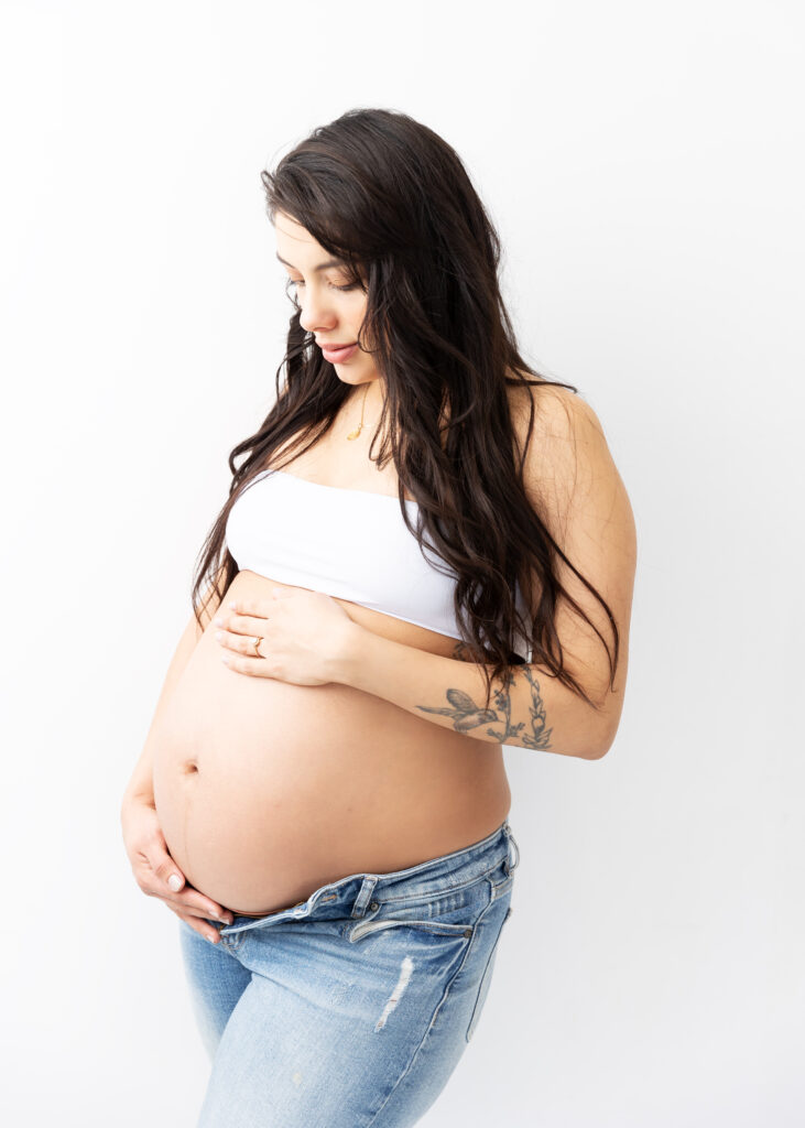 Pregnant woman in jeans and white top with tattoo cradling belly