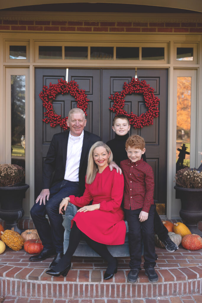 family sitting outside in front oh front door, mom in red dress sitting in chair, kids around her, deck the halls in-home holiday mini session