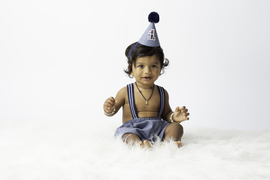 one year old boy sitting on white fur with hat and suspenders on