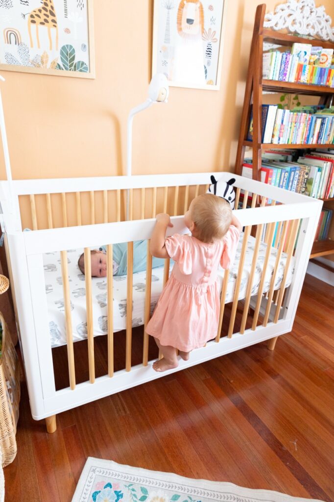 big sister climbing over crib to check on baby in colorful nursery in Oakley